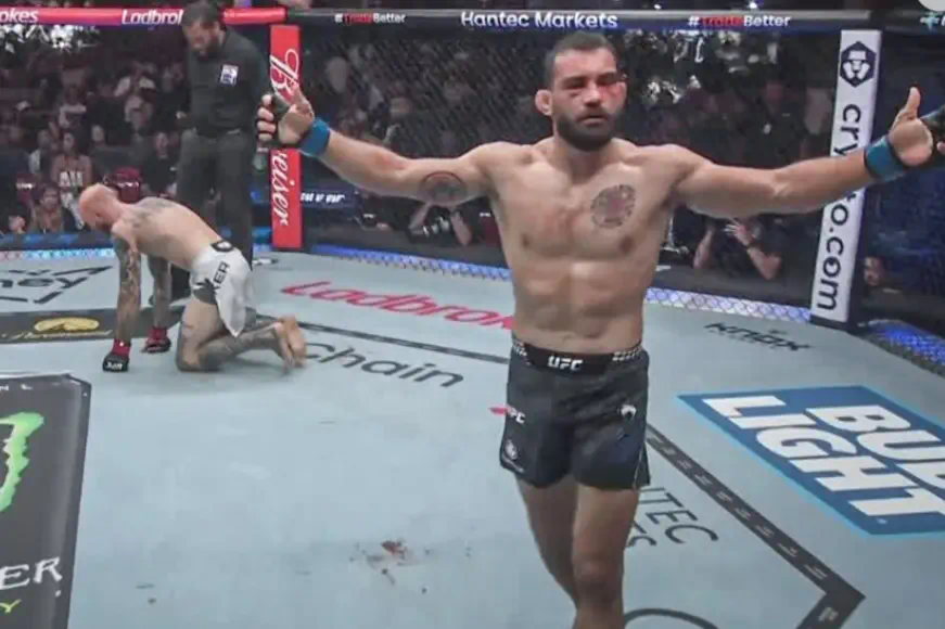 MMA fighter celebrating a victory inside the UFC octagon with arms extended, while the opponent kneels on the canvas after the fight.